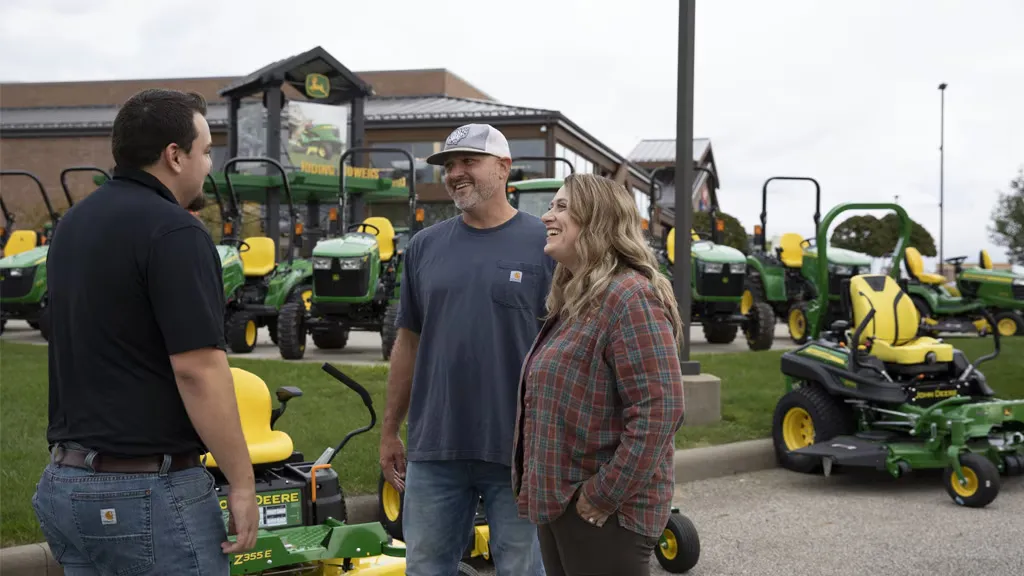 Salesperson talking with a couple in front of John Deere lawn equipment at a dealership