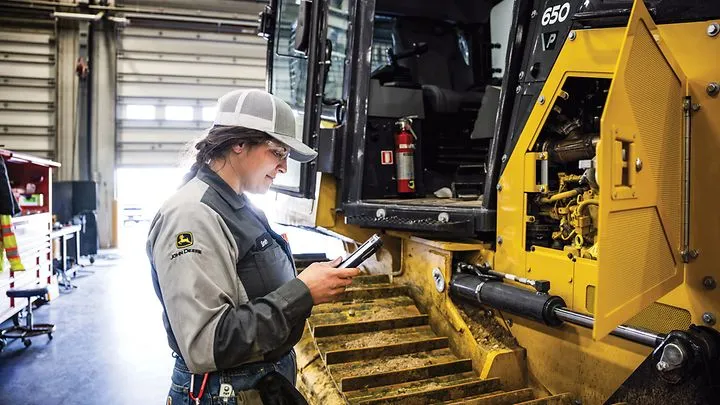 Technician using a tablet while inspecting open engine compartment of heavy equipment in a service bay
